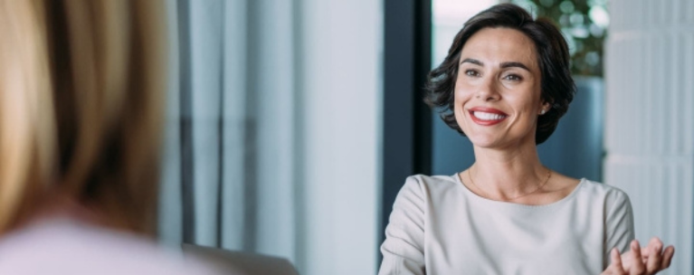 A professionally dressed woman smiling while talking to another woman turned away.